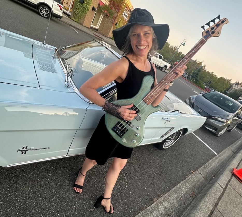 Kathy Johnson holding a bass guitar beside a classic car.