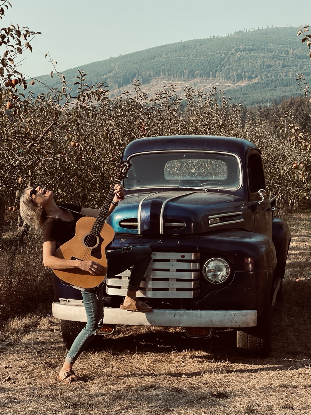 Kathy Johnson laughing with an acoustic guitar in front of a vintage truck in an orchard.