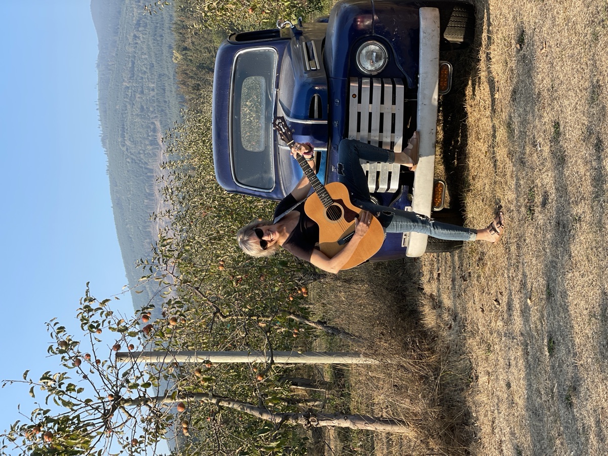 Kathy Johnson playing acoustic guitar near a vintage truck in an orchard.